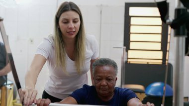 Kadın Pilates Koçu siyahi yaşlı bir kadına makine kullanmasında yardım ediyor, yaşlılık egzersizleri, esneklik ve güçlendirme.