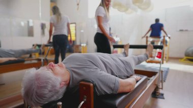 Pilates stüdyosunda egzersiz yapan yaşlı bir kadın. Yaşlı bir Kafkasyalı, Koç 'un rehberliğinde Makine' nin üzerine uzanmış ve vücudunu esnetiyordu.