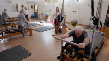 Grup oturumunda pilates makineleriyle egzersiz yapan yaşlılar. Bayan koç eğitmeni yaşlılara hareket ve vücut esnetme konusunda rehberlik ediyor. Yaşlılık egzersizi rutini