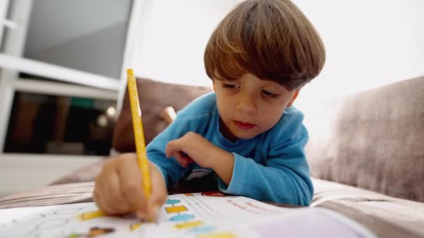 聚精会神的孩子用黄色钢笔在纸上画画 孩子们在家里躺在沙发上做学校活动 一个小男孩正挨着脸学习 — 图库视频影像