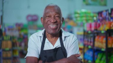 Afro-Amerikan üst düzey bir süpermarket çalışanının portresi önlük takıyor ve bakkal reyonunda kameraya gülümsüyor ve kollarını kavuşturuyordu.