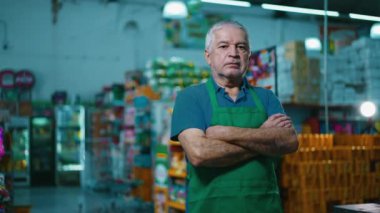 Market zincirinin içinde kolları çapraz duran ciddi bir küçük işletme sahibi. Önlük giyen yaşlı bir süpermarket müdürünün endişeli ifadesi
