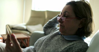 Kanepede oturmuş akıllı telefon ekranına bakan yaşlı bir kadın internetten video izliyor.