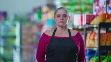 Endişeli orta yaşlı kadın bir süpermarket çalışanı endişeli bir ifadeyle koridorda duruyor, önlük giyiyor.
