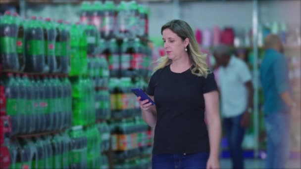 女消费者手握手机浏览产品 在超市苏打水饮料过道里走来走去 看食品杂货清单 妇女购物 描绘消费主义生活方式习惯 — 图库视频影像