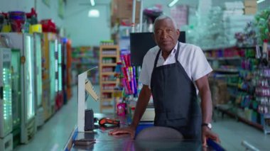 Süpermarketin siyah kıdemli bir çalışanı kasiyer portresinin önünde nötr bir ifadeyle önlük giyiyor. Ciddi Afro-Amerikan Market çalışanları