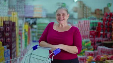 Süpermarkette alışveriş arabasıyla gülümseyen orta yaşlı kadın tüketici tüketim alışkanlıklarını tasvir ediyor
