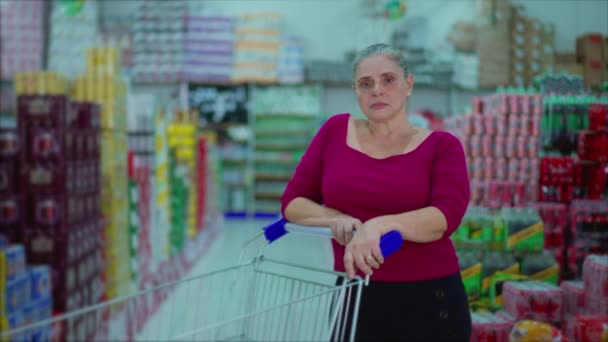 ショッピングカートや商品を背景にスーパーマーケットの通路の中に立っている一人の深刻な中年女性の買い物客 消費生活習慣を描いたカメラを見て — ストック動画