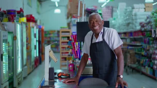 スーパーマーケットのレジに立っている幸せな黒人先輩がエプロンの制服を着たカメラで笑っている カリスマフレンドリーな食料品店のアフリカ系アメリカ人スタッフ — ストック動画