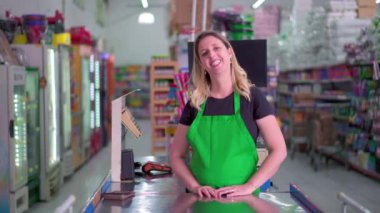Marketin mutlu bir bayan çalışanı kameraya gülümsüyor kasiyerin önünde duruyor yeşil önlükle gülümsüyor.