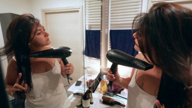 Saç kurutma makinesiyle banyo aynasının önünde saç kurutan bir kadın.