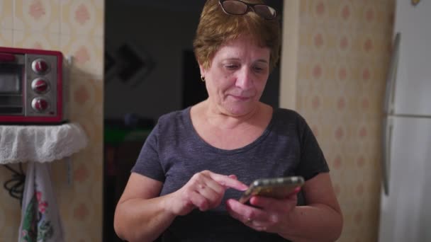 電話でメッセージを送っている間 自宅のキッチンに立っている携帯電話を使用するシニア女性 現代技術に携わる70代の高齢者 — ストック動画