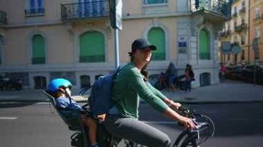 Bisikletli anne arka koltukta bebekle sokakta bisiklet sürüyor.