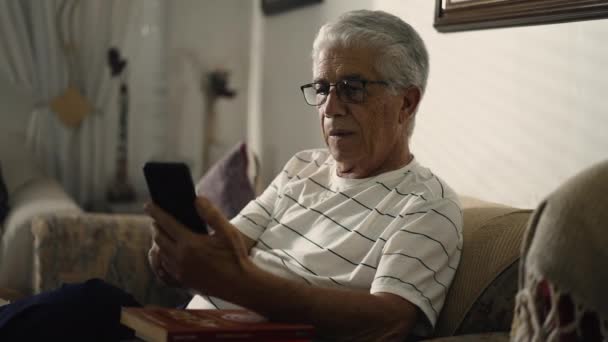 年配の男性は 懸念の表情で自宅のソファーに座って携帯電話デバイスを見つめ 困惑した感情でオンラインでビデオメディアを見ています — ストック動画