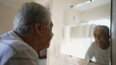 Yaşlı beyaz erkek, banyo aynasında kendi yansımasına bakıyor. Yaşlı, kır saçlı, yaşlılıktan pişmanlık ve üzüntü duyan yaşlı bir adam.