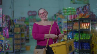 Marketin içinde duran mutlu bayan alışveriş sepeti. Markette gülümseyen orta yaşlı bir kadın tüketim yaşam tarzını tasvir ediyor