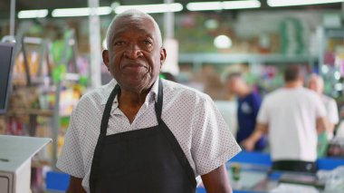 Süpermarketin siyah kıdemli çalışanlarının portresi kasiyerin önünde kameraya ciddi bir tarafsız ifadeyle bakıyor.