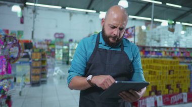 Marketin Brezilyalı çalışanı, süpermarket zincirinin içinde duran envanteri kontrol etmek için tablet kullanıyor, önlük takıyor ve işiyle meşgul.