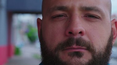 Ciddi bir sakallı erkek. Ciddi bir ifadeyle caddede kameraya bakıyor. Yakın plan portre yüzü.