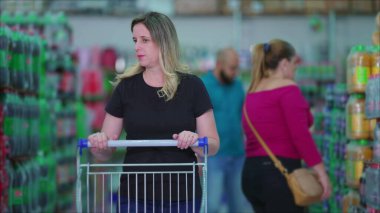 Market koridorunda alışveriş arabalarını iten kadın, arka planda tüketim alışkanlıklarını tasvir eden müşterilerle birlikte.