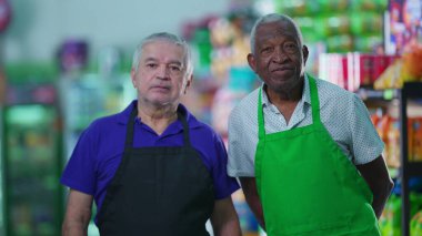 Süpermarketin iki kıdemli çalışanı tarafsız bir ifadeyle kameraya bakıyor. 70 'li yaşlarda önlük giyen yaşlı işçiler.