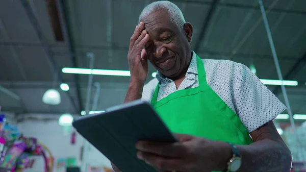 Frustrated African American Senior Employee Struggling with Tablet Device Inside Supermarket, Elderly Staff with Modern Technology