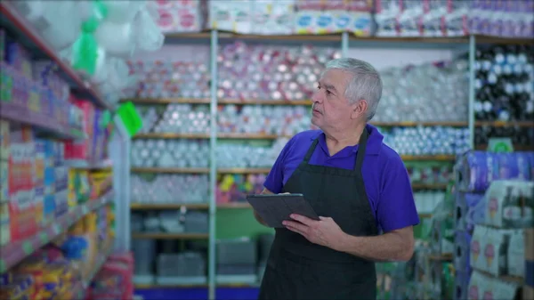 Senior manager of grocery store checking inventory of products on shelf with table device, male caucasian employee inspecting items at supermarket aisle