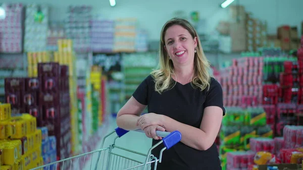 Female shopper standing at supermarket aisle with shopping cart smiling at camera. One caucasian woman consumer in mid 40s
