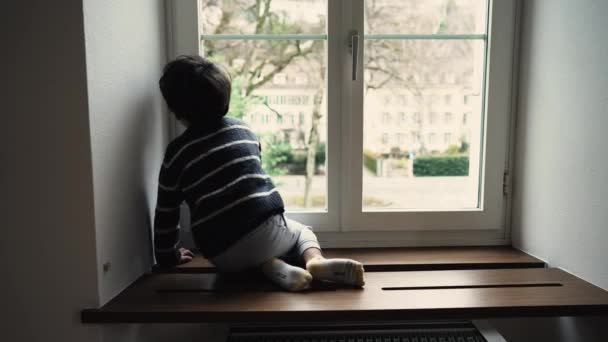 一个小男孩坐在窗边靠近玻璃的木架上 — 图库视频影像