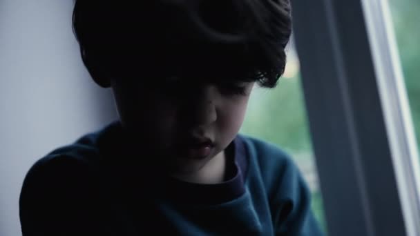 暗闇の中で窓に座っている悲しみの落ち込んだ子供 暗く暗いムードのメラノリックな小さな男の子は 屋内の窓で顔を閉じます — ストック動画