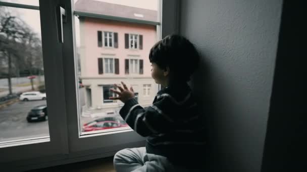 窓の外を眺める一人の退屈な子供 外に出たい小さな男の子 何もしない悲しい抑圧された子供 セカンドフロアの家から通りを眺める — ストック動画