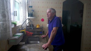 Düşünceli yaşlı bir adam mutfak lavabosunun yanında durup pencereden dışarı bakıyor. 70 'lerde derin derin düşünen yaşlı bir erkeğin ev hayatı sahnesi.