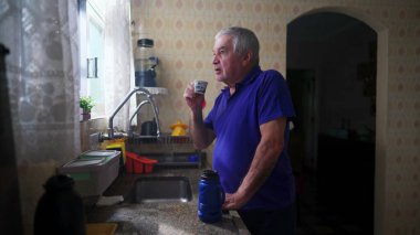 Son sınıf öğrencisi mutfak lavabosunun yanında durup kendine kahve ikram ediyor ve tefekkür ederek pencereden dışarı bakıyor.