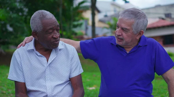 Two diverse senior friends sharing moment of camaraderie sitting on park bench, older caucasian friend putting hand over African American colelague shoulder in conversation