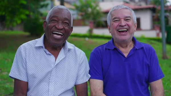 Two joyful Diverse senior friends laughing and smiling together on park bench outdoors. Happiness in old age retirement years. Portrait of Brazilian people