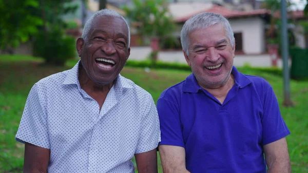 Two joyful Diverse senior friends laughing and smiling together on park bench outdoors. Happiness in old age retirement years. Portrait of Brazilian people