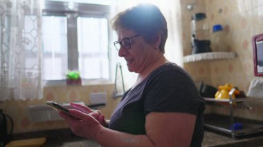 Neşeli Yaşlı Kadın Teknolojiye Bağlandı, Ev Mutfağında Cep Telefonu İçeriğini Okurken