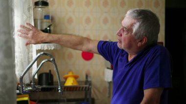 Son sınıf öğrencisi mutfak penceresinin yanında dikilip perdeden dışarı bakıyor evde tek başına manzarayı seyrediyor, yaşlı insan içeride güneş ışığını hissediyor.