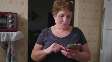 Ev mutfağında cep telefonu kullanan kıdemli bir kadın telefonla mesaj yolluyor. 70 'lerdeki yaşlı insan modern teknolojiyle meşgul.