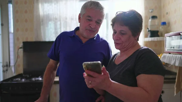 Senior wife showing cellphone screen to husband, elderly woman sharing online content to partner standing at home kitchen in casual domestic scene with real authentic people