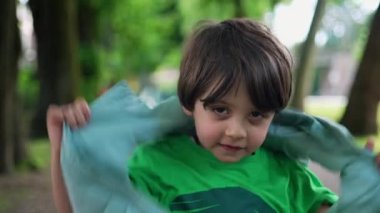 Kapüşonlu ceketli dört yaşındaki çocuk yeşil yolda dikiliyor. Moda giysiler giyen küçük bir çocuğun yakın plan yüzü.