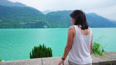 Düşünceli kadın arka planda dağlarla doğal göl manzarasının yanında duruyor. Dalgın bir kadın göl kenarına bakıyor, doğanın dinginliğinin ve huzurunun tadını çıkarıyor.