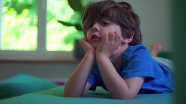 Huzursuz küçük çocuk evde kanepede uzanmış, elleri çenesinde, sıkılmış çocuk hareketsiz duramıyor ya da sakin olamıyor.