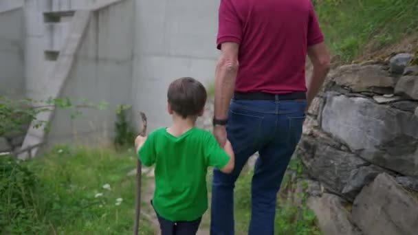 木の棒を握っている間 自然の道を歩いている祖父母の手を握る孫 祖父と孫のハイキング 家族の伴侶 — ストック動画