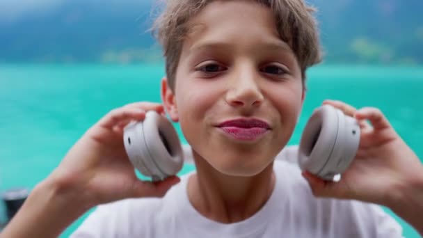 幸せな若い十代の少年がヘッドフォンを置く肖像画 オーディオブック ポッドキャスト または湖を見下ろす曲を聞いている10代の子供のクローズアップ顔 — ストック動画