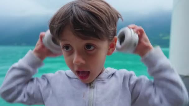 かわいい小さな男の子が 湖を背景に見下ろすヘッドフォンに乗っています イヤーヘッドホンに身に着けている幸せな子供の肖像 4歳の子供 — ストック動画