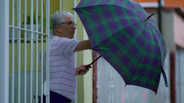 Olgun bir adam kötü havada şemsiye açar. Yaşlı beyaz adam yağmur yağarken evden çıkıp sokağa çıktı.