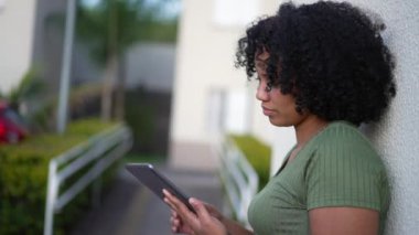 Açık havada tablet kullanan genç bir zenci kadın. Afro-Amerikalı yetişkin bir kız duvara yaslanmış ekranlara dokunuyor.