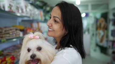 Evcil hayvan dükkanında duran bir kadın, küçük bir köpeği kollarında tutarken rafta duran ürünleri gözlemliyor.