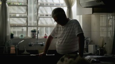 Dramatik, yalnız, yaşlı insan, mutfak penceresinin kenarında gölgelerde dikiliyor, başını eğip, endişe ve yalnızlık hissediyor.